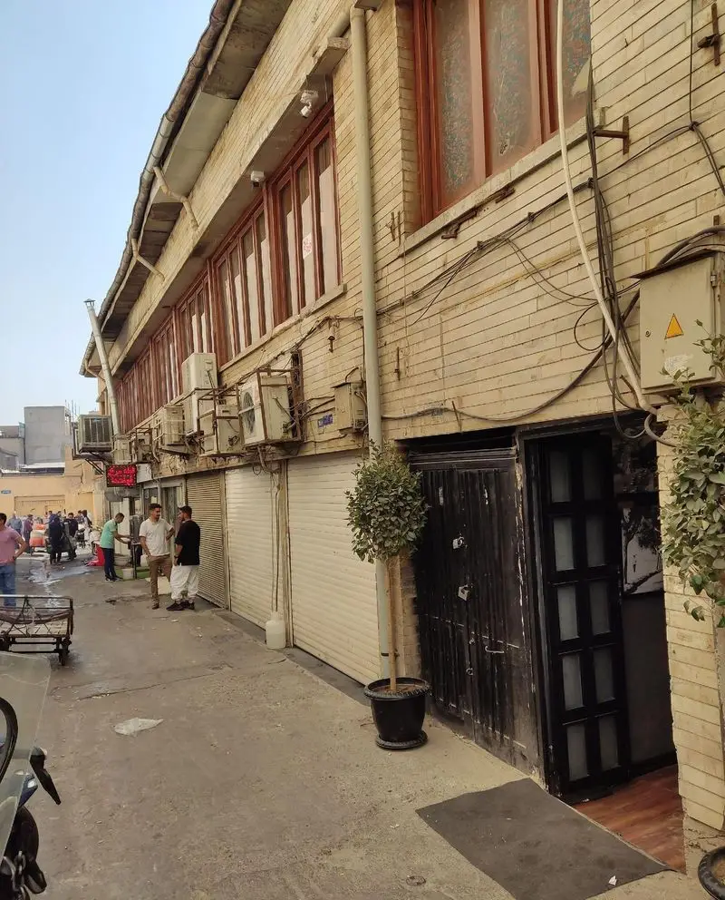 نمای بیرونی رستوران دیزی دنج طهرانی در یک کوچه قدیمی و شلوغ تهران، با درب چوبی مشکی، پنجره‌های آجری و فضای سنتی دلنشین.