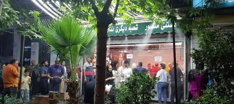 مغازه بستنی منصور شلوغ با سیستم مه پاش خنک کننده، مشتریان فراوان و درختان سبز در یک روز گرم تابستانی.