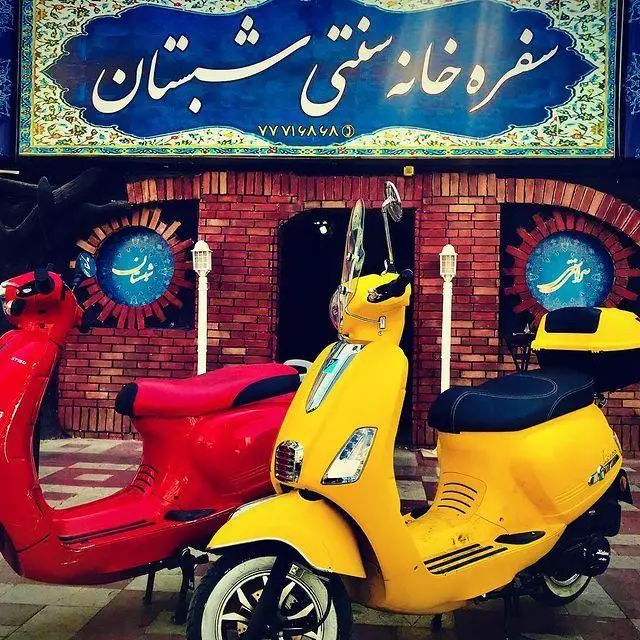 سفره خانه سنتی شبستان: ورودی رستوران با تابلوی بزرگ آبی رنگ و نوشته فارسی، به همراه دو اسکوتر کلاسیک شیک زرد و قرمز در مقابل بنای آجری.