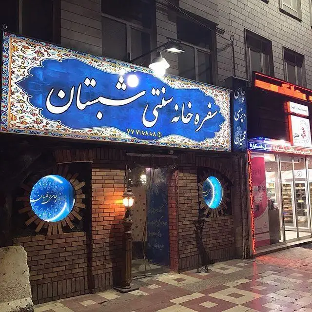 نمای شبانه سفره خانه سنتی شبستان با تابلوی آبی بزرگ، پنجره‌های دایره‌ای روشن و معماری آجری. مکانی برای غذای سنتی ایرانی.