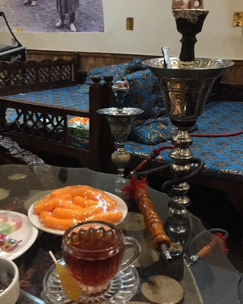 تصویری از فضای دلنشین سفره خانه سنتی زیر بازارچه شامل تخت‌های چوبی با روکش آبی، قلیان، چای داغ و شیرینی‌های سنتی.