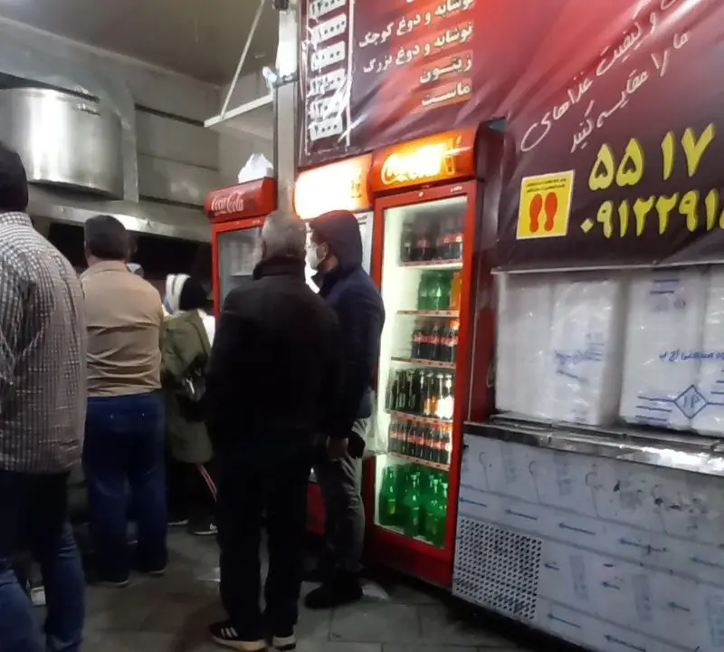 رستوران ارکیده خوش: مشتریان در حال بررسی منوی نوشیدنی‌ها، دوغ، زیتون و ماست، با شعار مقایسه کیفیت غذا. محیط شلوغ.