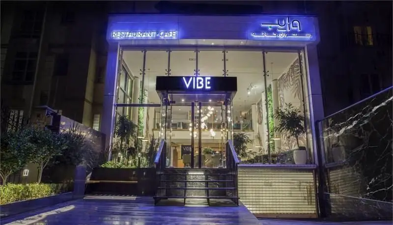 نمای بیرونی شیک و مدرن رستوران و کافه وایب در شب با نورپردازی آبی، ورودی شیشه‌ای جذاب و فضای سبز دلنشین.