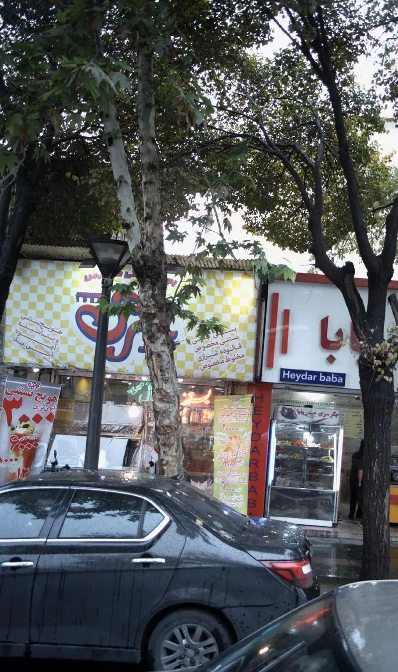 نمایی از مغازه فالوده و بستنی سنتی مخصوص جهرمی، با تابلوهای روشن بستنی، فالوده شیرازی و هویج بستنی، درختان و خودروها.