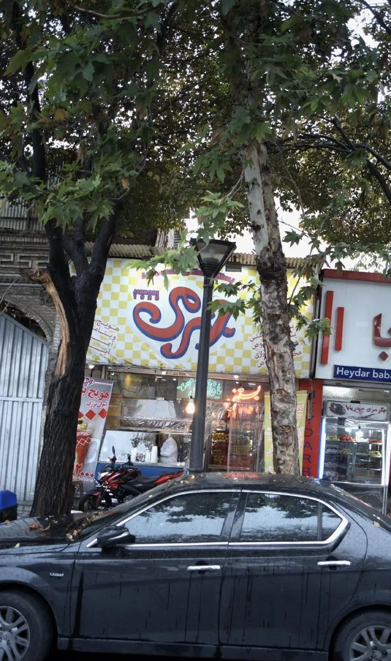 تصویری از فروشگاه فالوده و بستنی سنتی مخصوص جهرمی، با تابلوی نارنجی رنگ برزی روی زمینه شطرنجی و درختان بلند در اطراف.