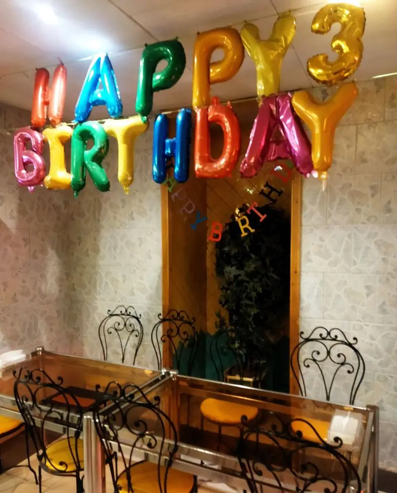 دکوراسیون تولد شاد و رنگارنگ کافی شاپ فرد یاس، شامل بادکنک‌های حروف "HAPPY BIRTHDAY" و میز و صندلی‌های شیک فرفورژه.