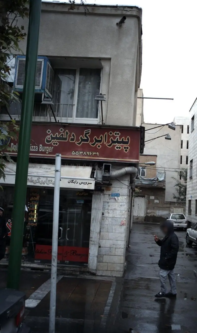 تصویری از نمای بیرونی فست فود پیتزا برگر دلفین در خیابان، با تابلوی اصلی، شماره تماس و اشاره به سرویس رایگان پیتزا و برگر.