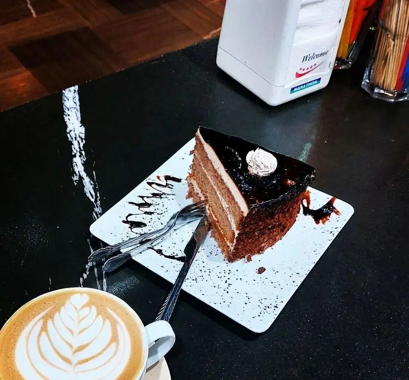 یک برش کیک شکلاتی سه لایه غرق در گاناش و خامه، همراه با قهوه لاته هنری در کافه رستوران کندو، انتخابی عالی برای لحظات شیرین.