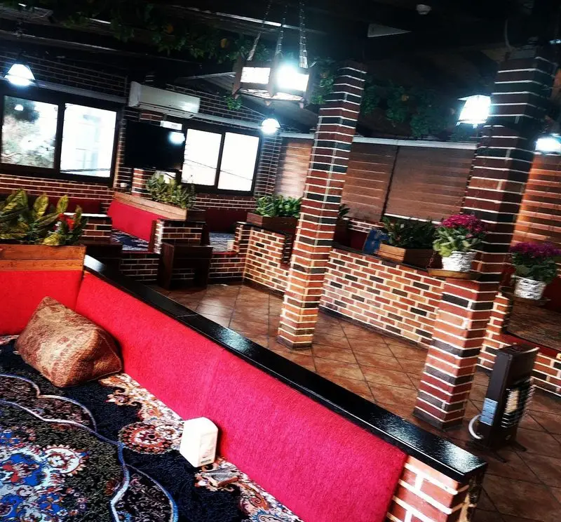 نمای دنج از کافه رستوران کندو با دیوارهای آجری، سکوهای نشیمن قرمز رنگ، فرش سنتی و گیاهان سبز برای تجربه دلپذیر.