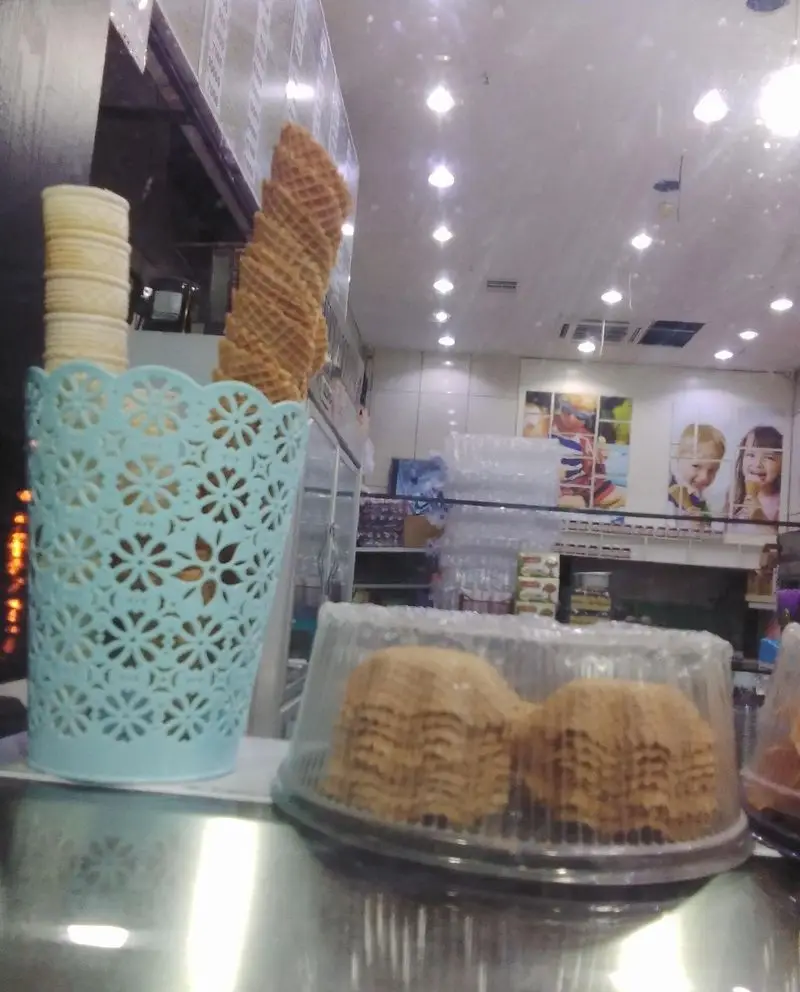 نمایی از قیف‌های بستنی کریسپی و کاغذی در بستنی فروشی ونیز، آماده سرو. محیط روشن با تصاویر شاد کودکان در پس‌زمینه جذاب.