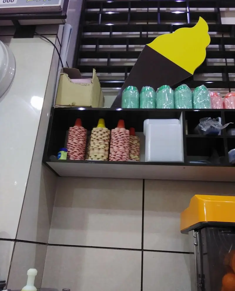 تصویری از فضای داخلی بستنی ونیز با قفسه‌های مرتب شامل لیوان‌های بستنی رنگی، ظروف یکبار مصرف، آبمیوه‌گیری و علامت بستنی قیفی.