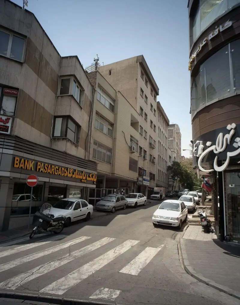 نمای شهری از کافه عربی امیران و بانک پاسارگاد در خیابان شلوغ تهران، با خودروها، موتور سیکلت و خط‌کشی عابر پیاده در روز.