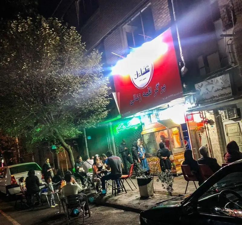 فست فود شقایق با تابلوی درخشان برگر لقمه ذغالی در شب، مشتریان در فضای باز خیابان در حال صرف غذا و تجربه محیط پرجنب‌وجوش.