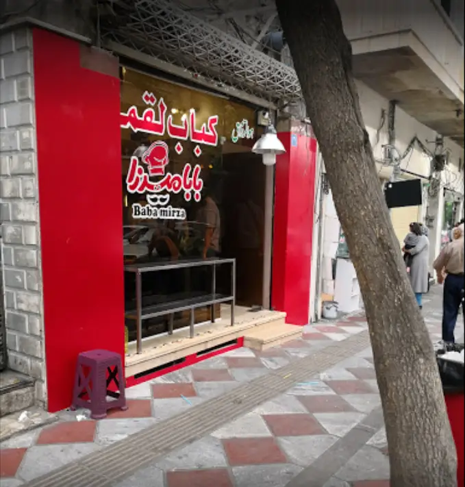 تصویر نمای بیرونی مغازه کباب لقمه "بابا میرزا" با تابلوی بزرگ و قرمز رنگ، ورودی شیشه ای و پیاده رو.