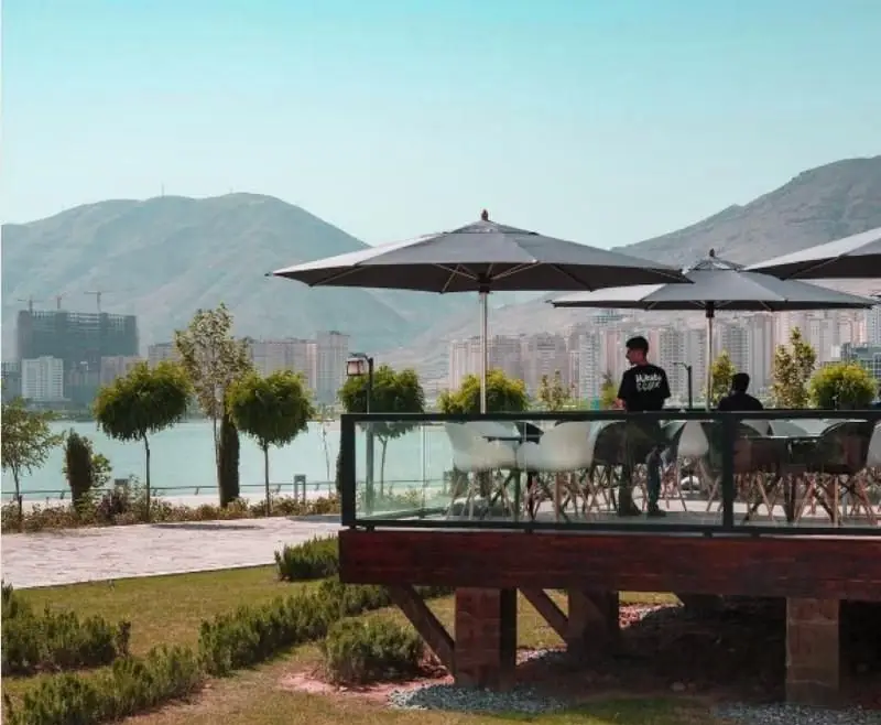 فضای باز دنج رستوران فرنگی کوبابا با میز و صندلی، چترهای بزرگ، منظره‌ای چشم‌نواز از دریاچه، شهر و کوهستان در پس‌زمینه.