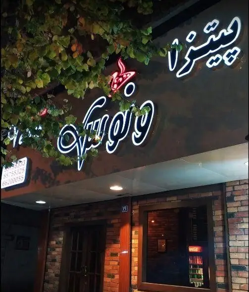 تابلوی نئون درخشان پیتزا ونوس در شب، با نمای آجری ساختمان، شاخ و برگ سبز و درب ورودی، به همراه پلاک و شماره تماس‌های مغازه.