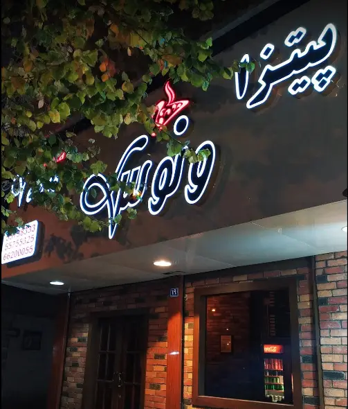 تصویری از نمای شبانه رستوران فست فود پیتزا ونوس با تابلو نئون روشن جذاب و لوگوی قرمز رنگ، همراه با برگ‌های سبز و نمای آجری.