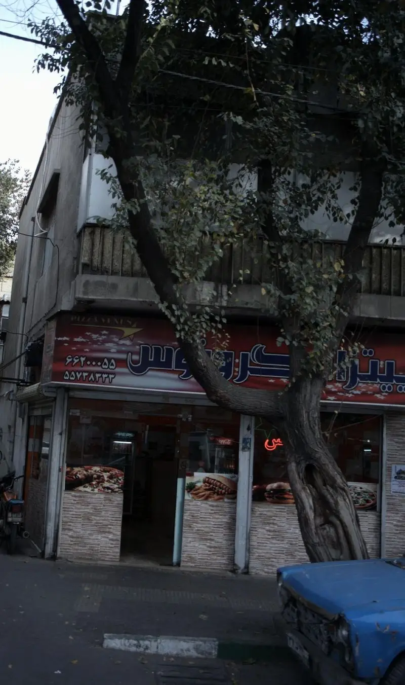 نمای بیرونی پیتزا فروشی ونوس (Pizza Venus) در خیابان، با تابلوی قرمز رنگ بزرگ، شماره‌های تماس، و درخت بلند جلوی مغازه فست فود.