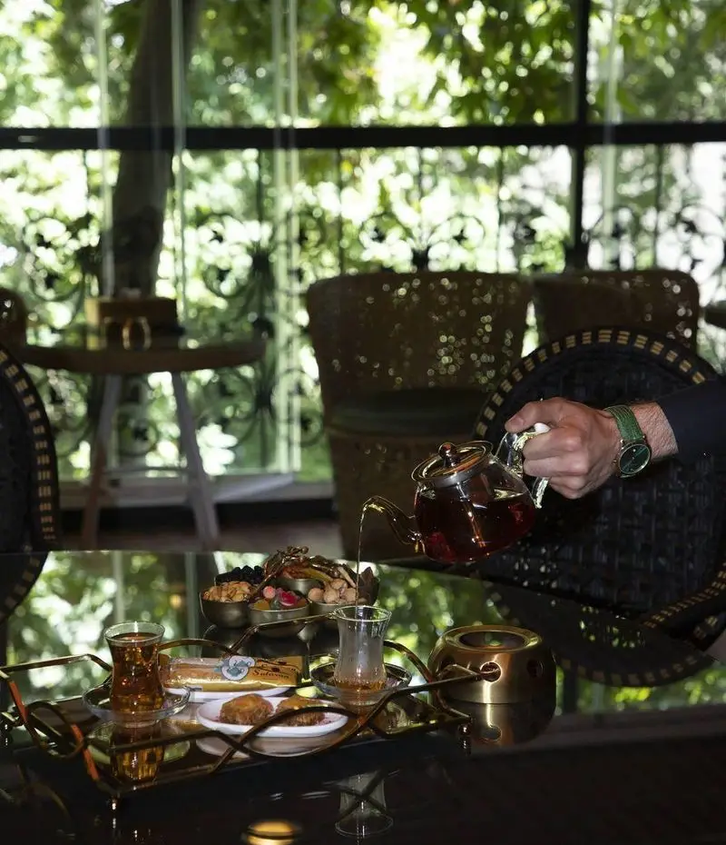 کافه سالار: ریختن چای خوش‌رنگ در استکان کنار سینی طلایی حاوی دسر، شیرینی، آجیل و میوه‌های خشک در محیطی آرام با چشم‌انداز سبز.