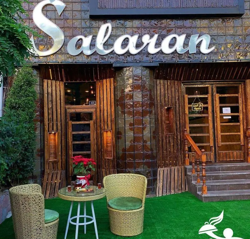 نمای بیرونی کافه سالار با دکوراسیون چوبی و کاشی‌های براق، فضای باز با چمن مصنوعی و میز و صندلی حصیری برای استراحت و نوشیدنی.