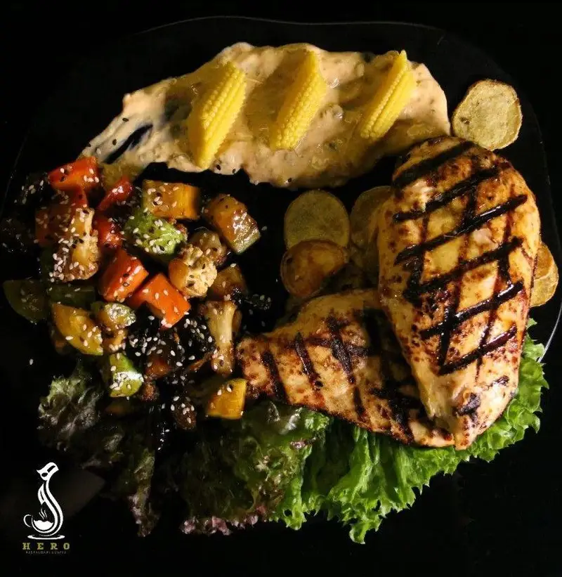 سینه مرغ گریل شده با سبزیجات متنوع، ذرت کوچک، سس خامه‌ای و کاهو تازه، غذایی سالم و لذیذ در کافه رستوران هرو.