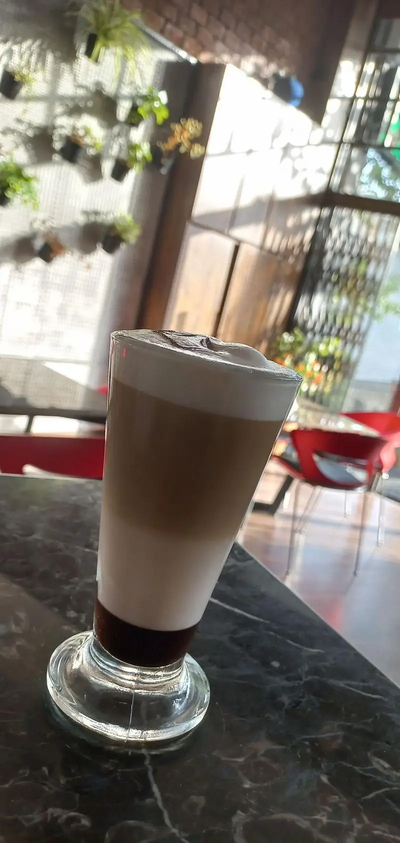 یک لیوان لاته خوش‌طعم و چند لایه در کافه رستوران ورسی، روی میز مرمری در فضایی روشن و دلنشین با گیاهان و دکور مدرن.
