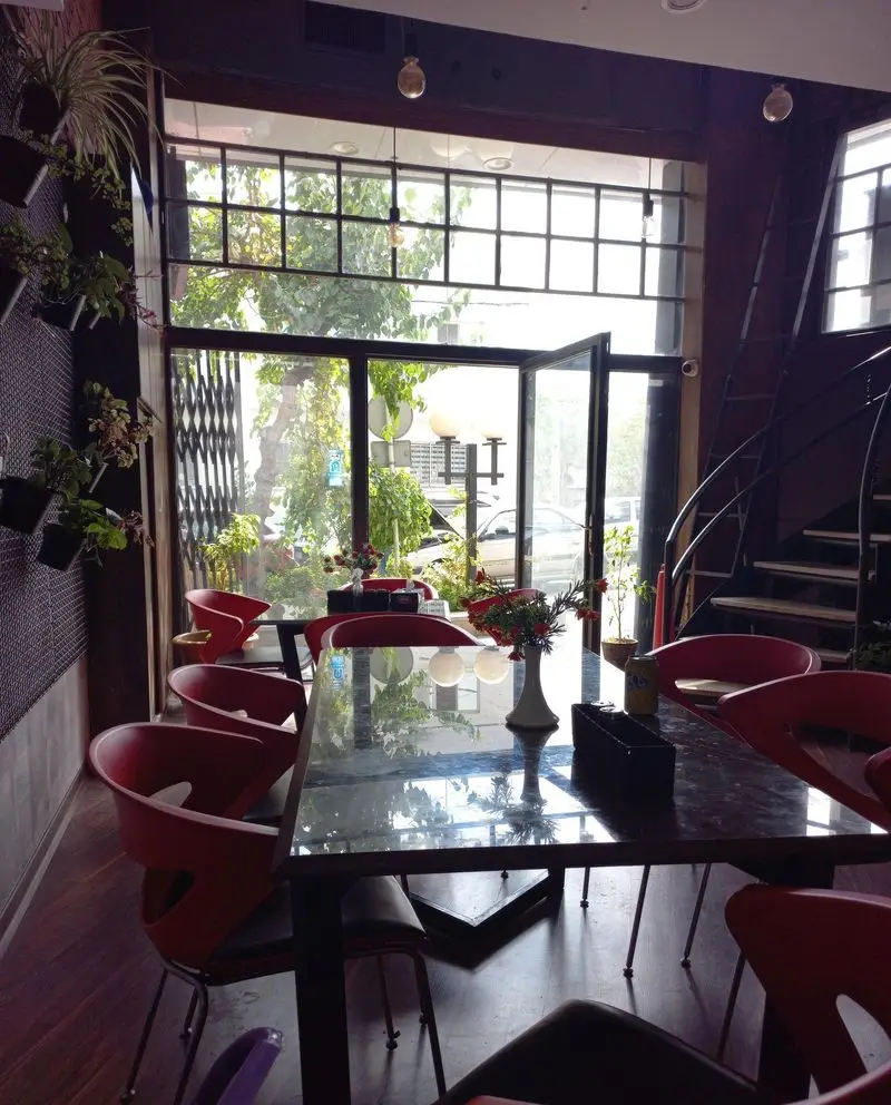 فضای داخلی شیک کافه رستوران ورسی با صندلی‌های قرمز، پنجره بزرگ، گیاهان، میزهای مدرن و پله مارپیچ.