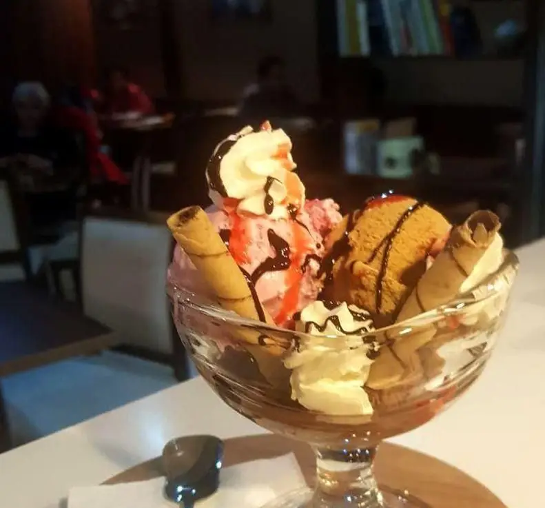 بستنی لیوانی لذیذ کافه نتو با اسکوپ‌های توت‌فرنگی و کارامل، تزئین شده با خامه، سس شکلات و بیسکویت ویفر، در فضای دلنشین کافه