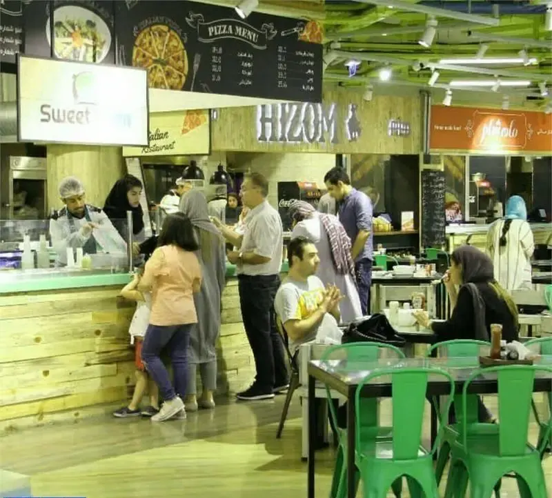 فودکورت گورمه مارکت مزرعه ماهشام: مشتریان در حال سفارش غذا، کارکنان مشغول، با غرفه‌های پیتزا، ایتالیایی و میزهای سبز رنگ