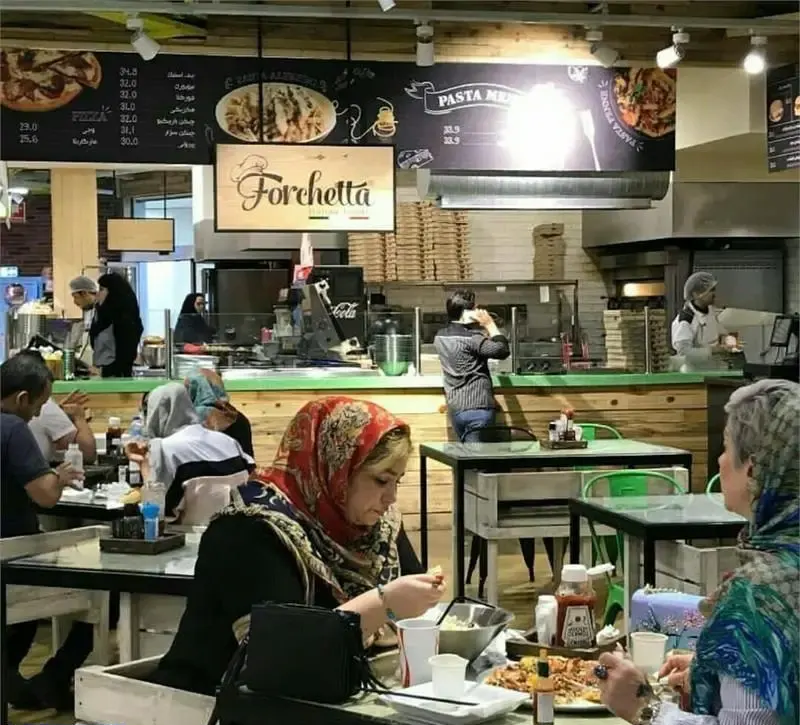 فضای داخلی رستوران گورمه مارکت مزرعه ماهشام با مشتریان در حال صرف غذا و کارکنان در آشپزخانه آماده سازی پیتزا و پاستا.