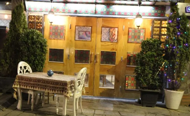 رستوران سنتی علی بابا: نمای بیرونی در شب، فضای باز با میز و صندلی و رومیزی طرح‌دار، درب‌های چوبی با تزئینات سنتی و نورپردازی گرم.
