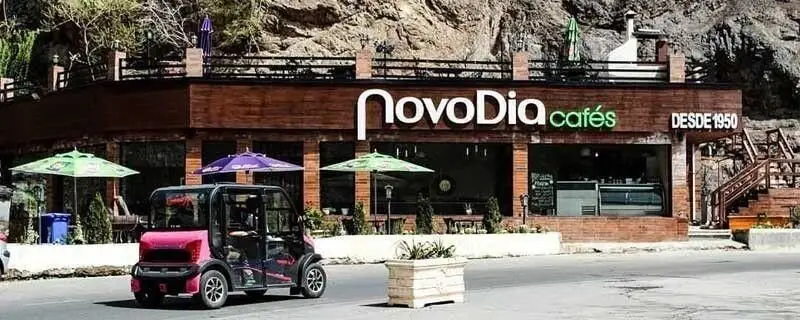 کافه نوودیا: نمای بیرونی جذاب با دکوراسیون چوبی و چترهای رنگی، فضای باز دلپذیر برای قهوه، با پس‌زمینه کوهستانی (از ۱۹۵۰).