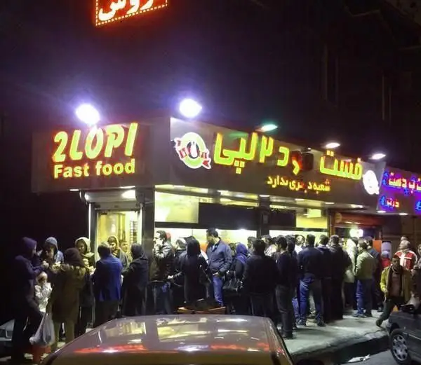 فست فود دو لپی: نمای شبانه شلوغ با صف مشتریان، تابلوهای نئونی روشن و فضای پر جنب و جوش برای فلافل و غذای فوری.