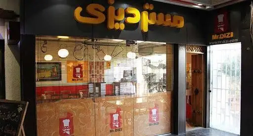 ورودی رستوران مستر دیزی با تابلو زرد رنگ برجسته، شیشه‌های طرح‌دار با لوگوی سرآشپز و آدرس وبسایت mrdizi.com، محیطی سنتی برای دیزی.
