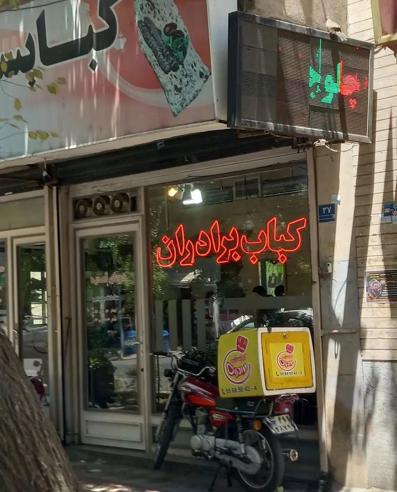 نمای سردر رستوران کباب برادران، با تابلوی کباب، نئون روشن کباب برادران و موتورسیکلت بیرون‌بر آن.