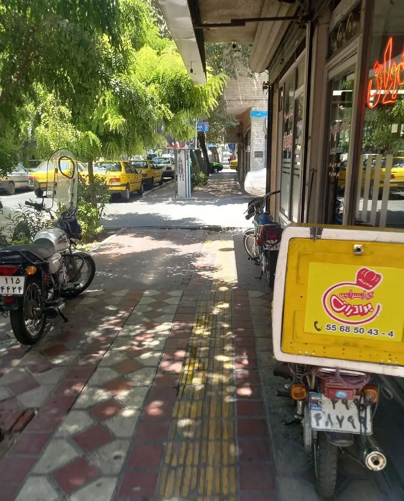 نمای خیابان از کبابسرای برادران با موتورسیکلت دلیوری، پیاده‌رو، درختان سبز و تاکسی‌های زرد در یک روز آفتابی.