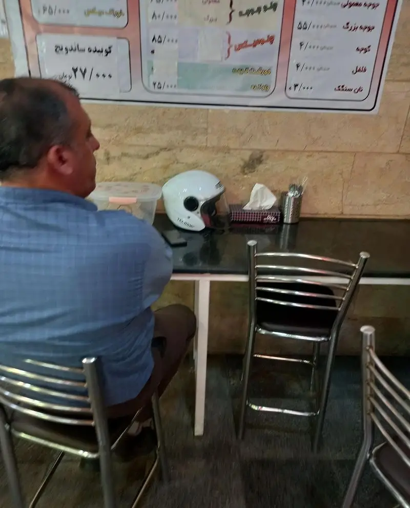 نمایی از یک مشتری در رستوران برادران، در حال انتخاب غذا از منوی نصب شده روی دیوار. کلاه ایمنی و دستمال کاغذی روی میز.