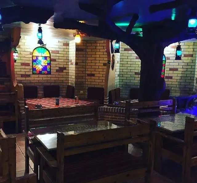 نمایی از فضای داخلی سنتی سفره خانه کاج با دیوارهای آجری، مبلمان چوبی، پنجره‌های رنگی، فانوس و ستون درختی بزرگ.