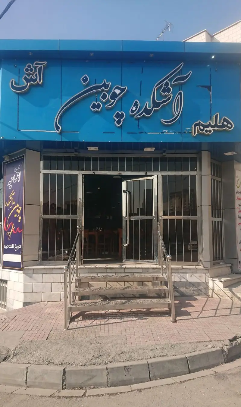 نمای بیرونی آشکده چوبین: رستوران سرو کننده آش و حلیم، با تابلوی آبی رنگ برجسته و درب ورودی شیشه‌ای.