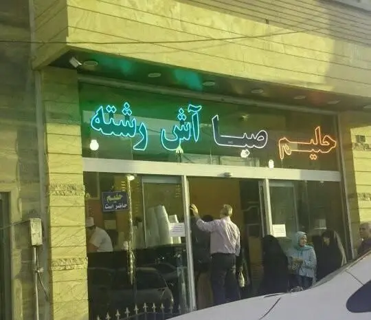 عکس نمای بیرونی آش و حلیم پزی حلیم صبا با تابلو نئون درخشان آش رشته و حلیم، مشتریان در حال خرید غذای سنتی و گرم ایرانی.