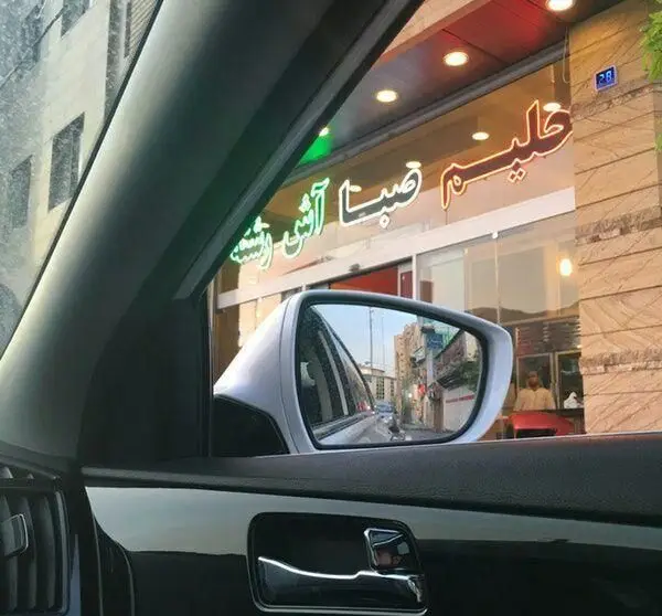 نمای آش و حلیم پزی حلیم صبا از داخل خودرو، با تابلوهای نئون درخشان "آش و حلیم" و "حلیم صبا" و بازتاب خیابان در آینه.