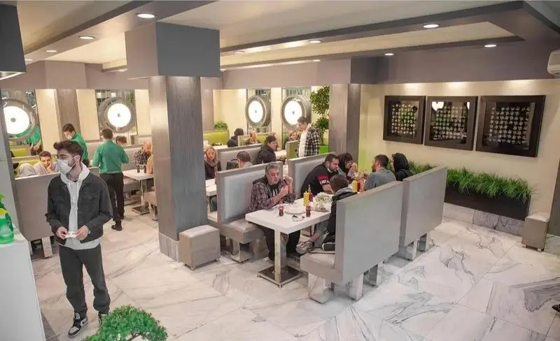 فضای داخلی بزرگ و مدرن پیتزا چمن با مشتریانی در حال صرف غذا و نوشیدنی. میز و صندلی‌های شیک، کفپوش مرمر و محیطی دلنشین برای فست فود.
