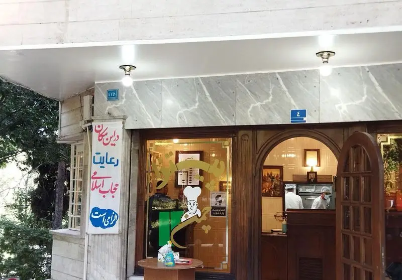 نمای بیرونی کسب و کار پیتزا چمن، با درب‌های چوبی سنتی، تابلوی رعایت حجاب اسلامی و لوگوی سرآشپز بر شیشه. دو آشپز داخل مشغول کارند.