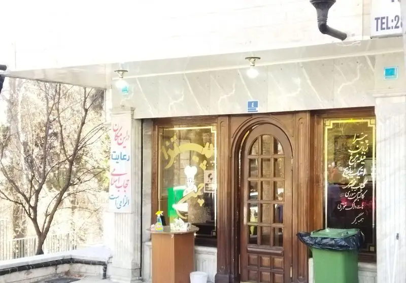 تصویری از نمای بیرونی پیتزا چمن، رستوران فست فود با در چوبی و ویترین منوی غذا. تابلوی الزامی بودن حجاب و لوگوی سرآشپز در ورودی.