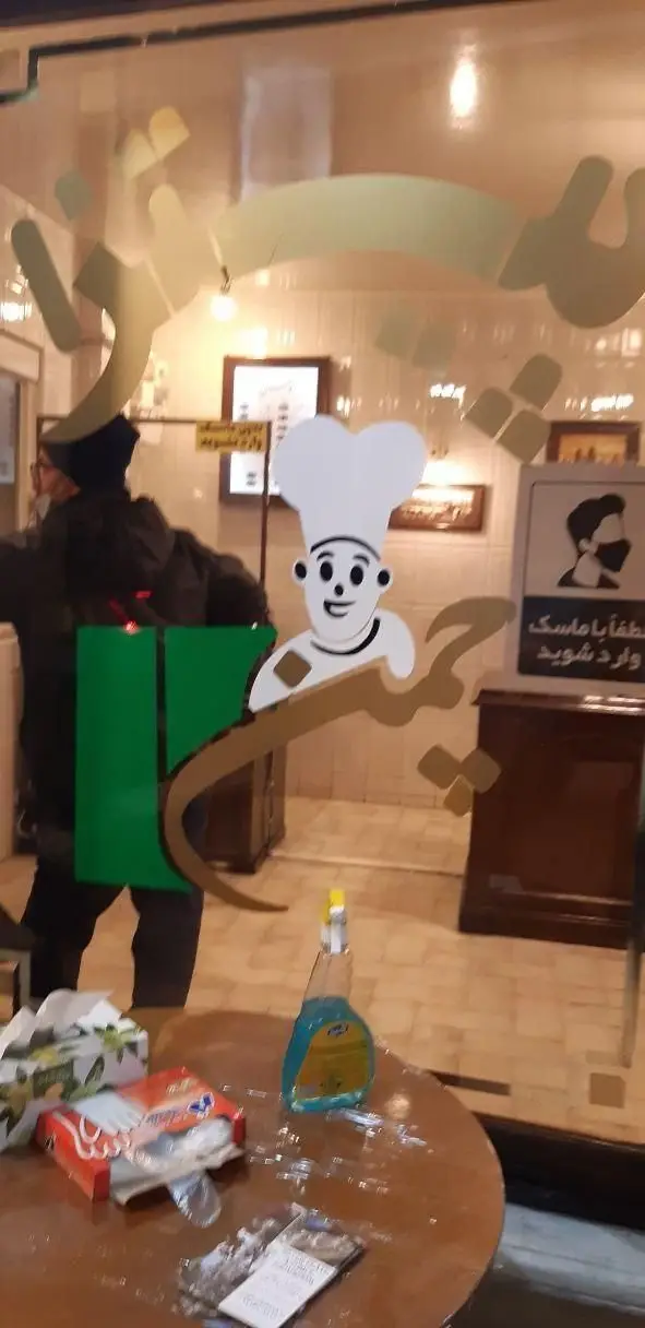پیتزا چمن: نمای داخلی ورودی رستوران با لوگوی جذاب سرآشپز و تابلوی رعایت پروتکل‌های بهداشتی (ماسک).