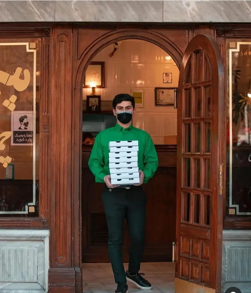 پیک پیتزا چمن با لباس فرم سبز و ماسک محافظ، هفت جعبه پیتزا را از درب چوبی رستوران برای تحویل سریع سفارش مشتریان حمل می‌کند.