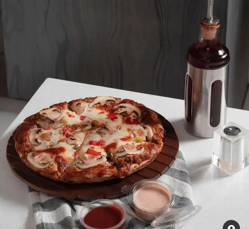 پیتزا قارچ چمن با پنیر فراوان و فلفل دلمه‌ای رنگی، سرو شده روی تخته چوبی با سس‌های مخصوص و ظرف روغن برای تجربه‌ای لذیذ.