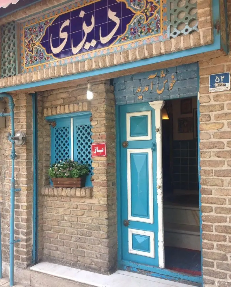 نمای بیرونی رستوران سنتی دیزی با تابلوی آبی کاشی‌کاری شده، درب ورودی فیروزه‌ای، پنجره گلدان‌دار و تابلوی باز، آماده پذیرایی است.