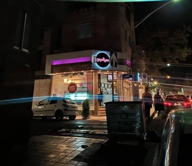 تصویری از نمای شبانه و جذاب قنادی سینامون با تابلوهای نورانی، نورپردازی بنفش و محیط پرتردد شهری، در شبی زیبا.