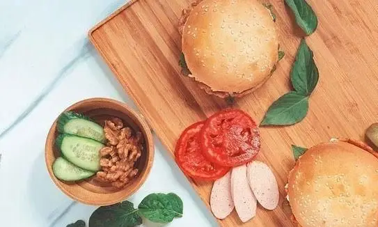 برگرهای تازه و خوشمزه از پیتزا پرپروک با نان کنجدی، گوجه و سبزیجات معطر، همراه با مخلفات سالم شامل خیار و گردو.